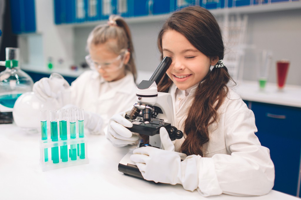 Two little kids in lab coat learning chemistry in school laboratory. Young scientists in protective glasses making experiment in lab or chemical cabinet. Looking through the microscope